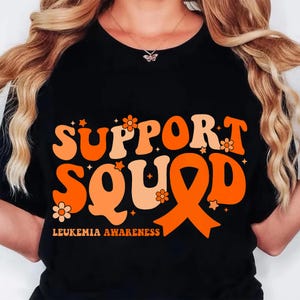 May include: Black t-shirt with the words "SUPPORT SQUAD" in orange and peach-colored retro font. The design includes an orange awareness ribbon and the text "LEUKEMIA AWARENESS" at the bottom. Small flowers and stars are scattered around the text.