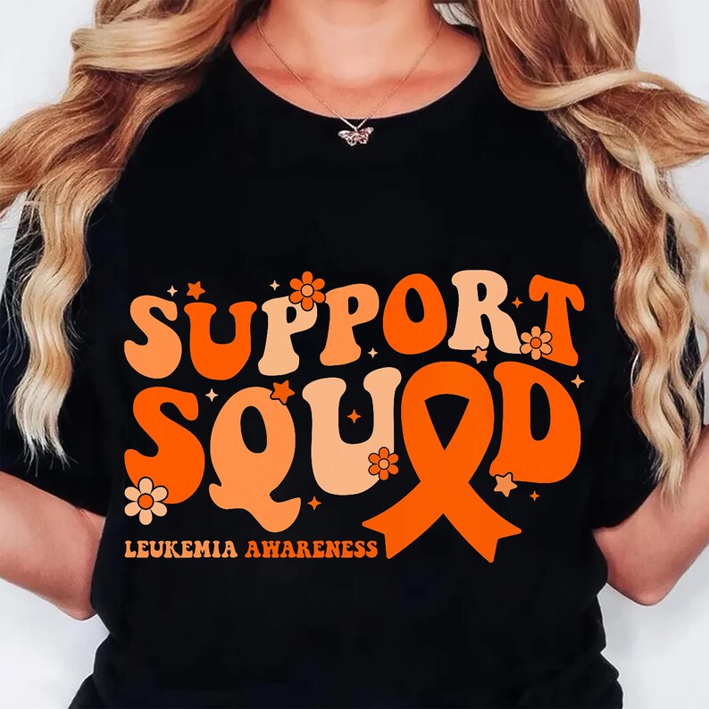 May include: Black t-shirt with the words "SUPPORT SQUAD" in orange and peach-colored retro font. The design includes an orange awareness ribbon and the text "LEUKEMIA AWARENESS" at the bottom. Small flowers and stars are scattered around the text.