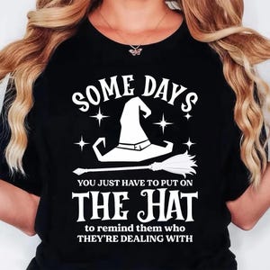 May include: Black t-shirt with a white graphic of a witch's hat and broom, with the text "SOME DAYS YOU JUST HAVE TO PUT ON THE HAT to remind them who THEY'RE DEALING WITH". A Halloween-themed graphic tee.