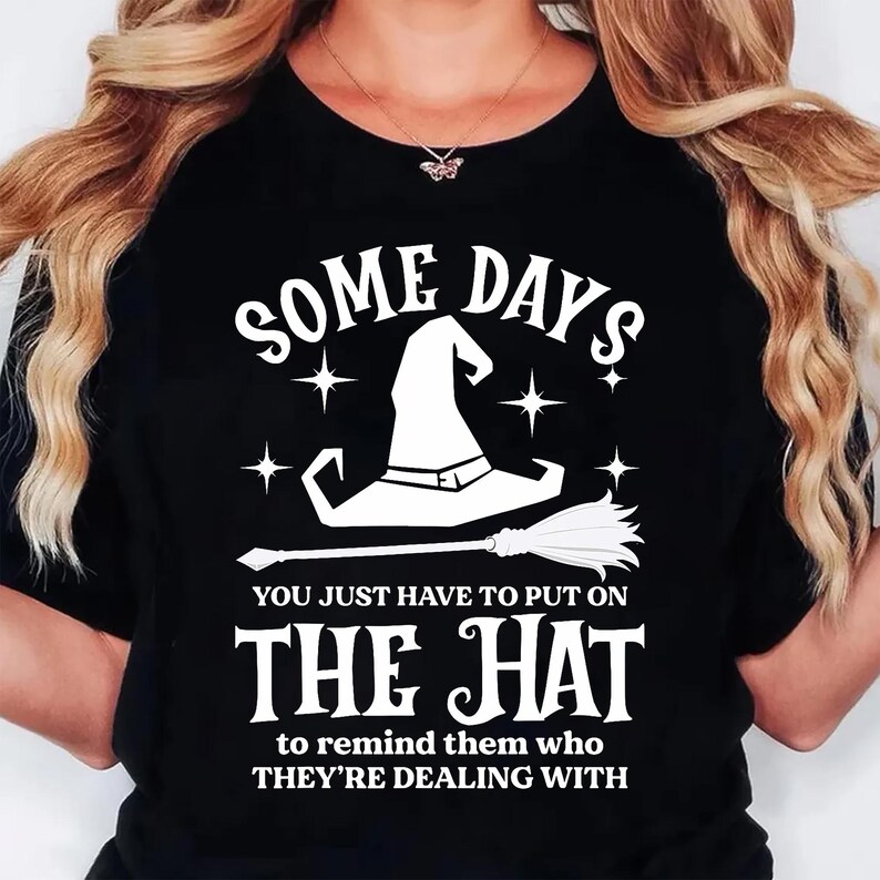 May include: Black t-shirt with a white graphic of a witch's hat and broom, with the text "SOME DAYS YOU JUST HAVE TO PUT ON THE HAT to remind them who THEY'RE DEALING WITH". A Halloween-themed graphic tee.