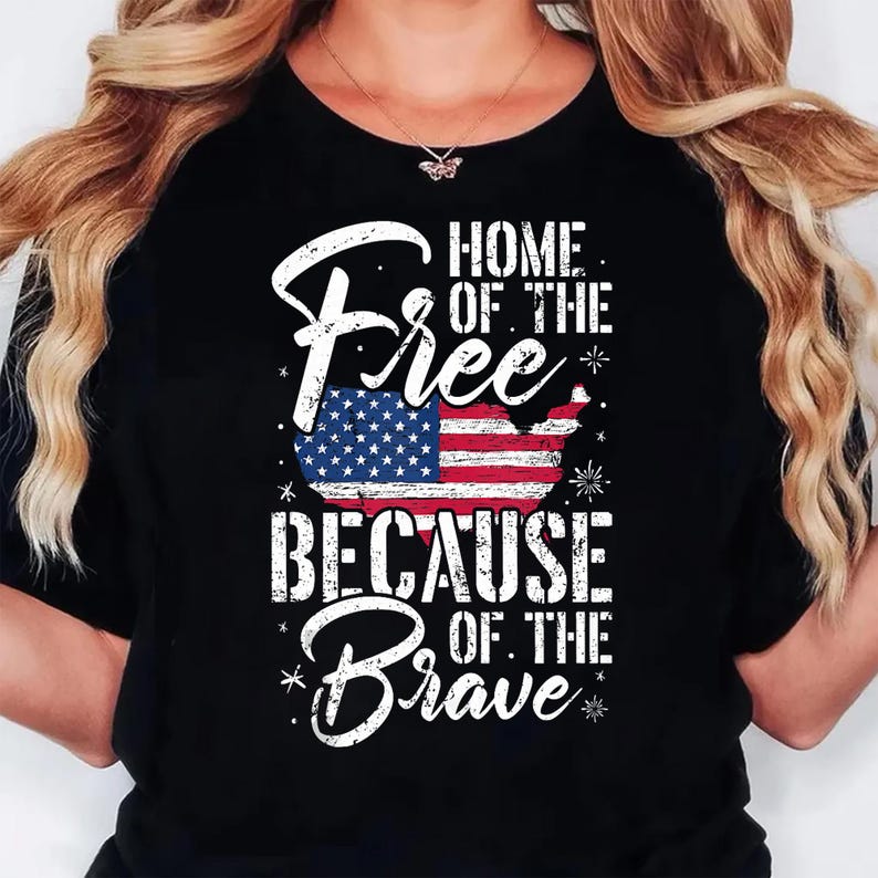 May include: A black t-shirt featuring a distressed white graphic with the text Home of the Free Because of the Brave. The design includes a stylized map of the United States filled with the American flag pattern, accented by small star and firework icons.