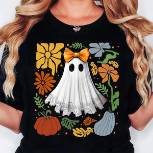 May include: Black t-shirt featuring a white ghost with an orange bow, surrounded by orange, yellow, and gray floral and pumpkin designs. The ghost has black oval eyes and a lace trim. A fun Halloween-themed graphic tee.