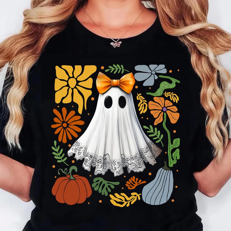 May include: Black t-shirt featuring a white ghost with an orange bow, surrounded by orange, yellow, and gray floral and pumpkin designs. The ghost has black oval eyes and a lace trim. A fun Halloween-themed graphic tee.