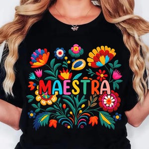 May include: Black t-shirt with a colorful floral embroidery design and the word "MAESTRA" in vibrant letters. The embroidery features flowers in shades of red, yellow, blue, and pink, with green leaves and stems.