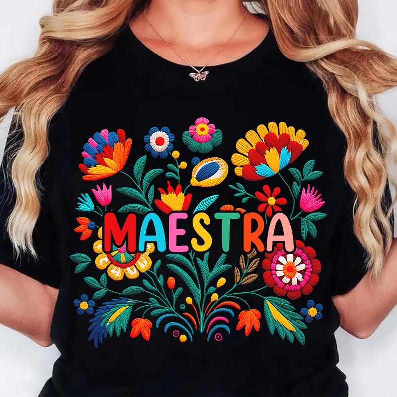 May include: Black t-shirt with a colorful floral embroidery design and the word "MAESTRA" in vibrant letters. The embroidery features flowers in shades of red, yellow, blue, and pink, with green leaves and stems.