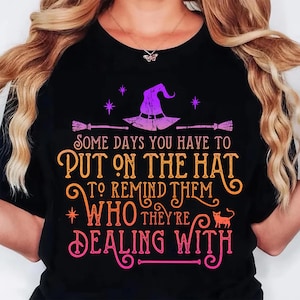 May include: Black t-shirt with a Halloween-themed graphic. The design features a witch's hat, broomsticks, stars, and the text "SOME DAYS YOU HAVE TO PUT ON THE HAT TO REMIND THEM WHO THEY'RE DEALING WITH."