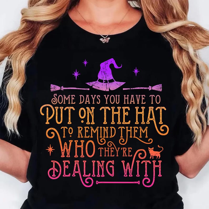 May include: Black t-shirt with a Halloween-themed graphic. The design features a witch's hat, broomsticks, stars, and the text "SOME DAYS YOU HAVE TO PUT ON THE HAT TO REMIND THEM WHO THEY'RE DEALING WITH."