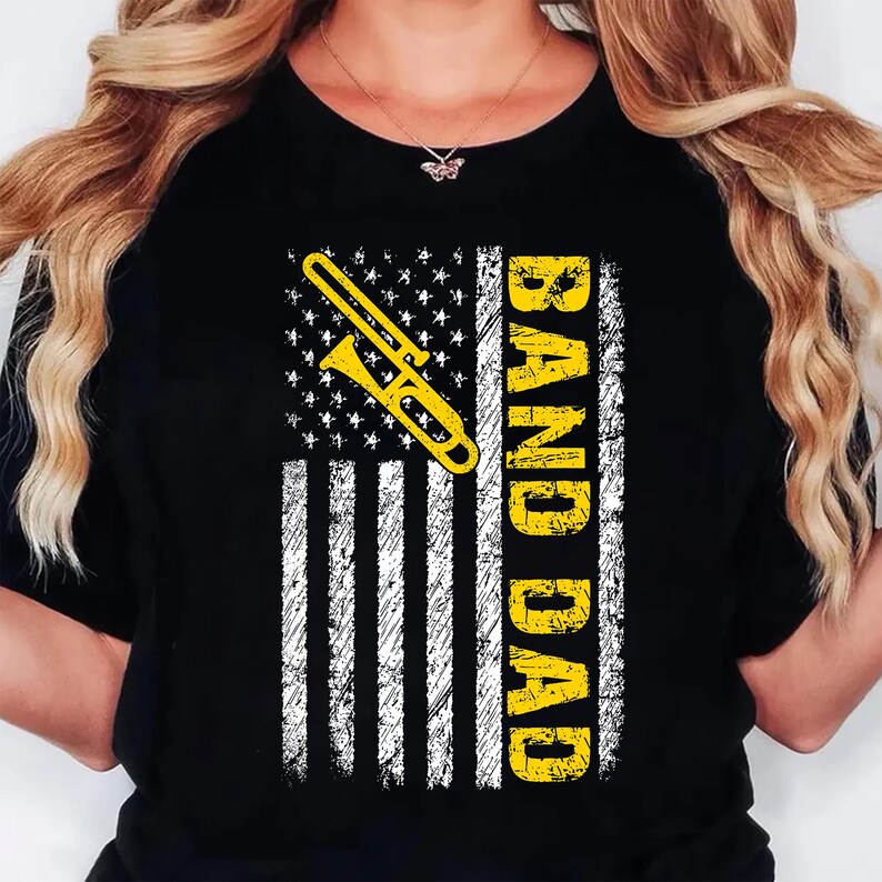 K&ouml;nnte beinhalten: Schwarzes T-Shirt mit einer gelben Posaunen-Grafik &uuml;ber einem abgenutzten amerikanischen Flaggen-Design. Die Worte "BAND DAD" sind vertikal in Gelb gedruckt. Ein Musik-Themen-Bekleidungsartikel.
