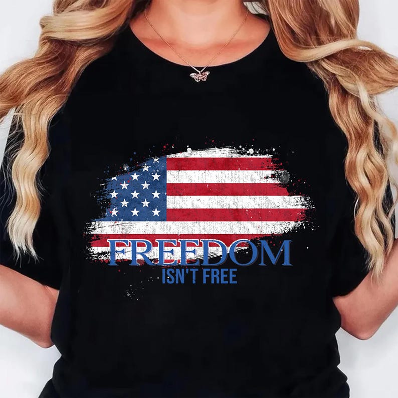 May include: A black t-shirt featuring a distressed American flag graphic across the chest. The design includes the text FREEDOM in large blue letters and the words ISN'T FREE in smaller blue letters directly underneath the flag graphic.