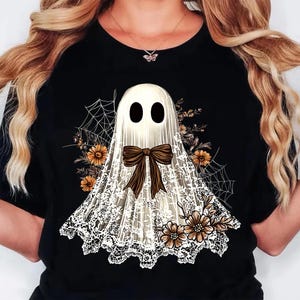 May include: Black t-shirt featuring a white ghost design with a brown bow, lace detailing, and floral accents. The ghost has two black oval eyes. Spiderwebs and flowers add to the Halloween theme.