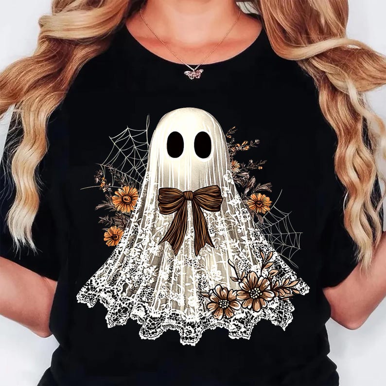 May include: Black t-shirt featuring a white ghost design with a brown bow, lace detailing, and floral accents. The ghost has two black oval eyes. Spiderwebs and flowers add to the Halloween theme.