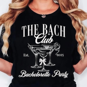 May include: Black t-shirt with white text and a cocktail graphic. The text reads "THE BACH Club" above a graphic of three martini glasses and the words "Bachelorette Party". The year "2025" is also printed on the shirt.