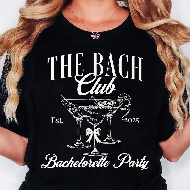 May include: Black t-shirt with white text and a cocktail graphic. The text reads "THE BACH Club" above a graphic of three martini glasses and the words "Bachelorette Party". The year "2025" is also printed on the shirt.