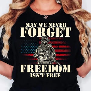 May include: A black t-shirt featuring a graphic of a soldier in tactical gear holding a rifle in front of a distressed American flag. The text reads MAY WE NEVER FORGET and FREEDOM ISN'T FREE in a bold, light-colored, stencil-style font.