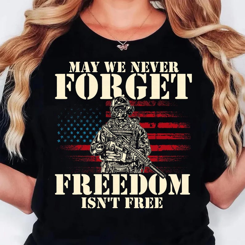 May include: A black t-shirt featuring a graphic of a soldier in tactical gear holding a rifle in front of a distressed American flag. The text reads MAY WE NEVER FORGET and FREEDOM ISN'T FREE in a bold, light-colored, stencil-style font.