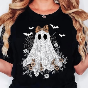 May include: Black t-shirt featuring a white ghost design with a brown bow, surrounded by bats, flowers, and a spiderweb. The ghost is made of lace. A Halloween-themed graphic tee.