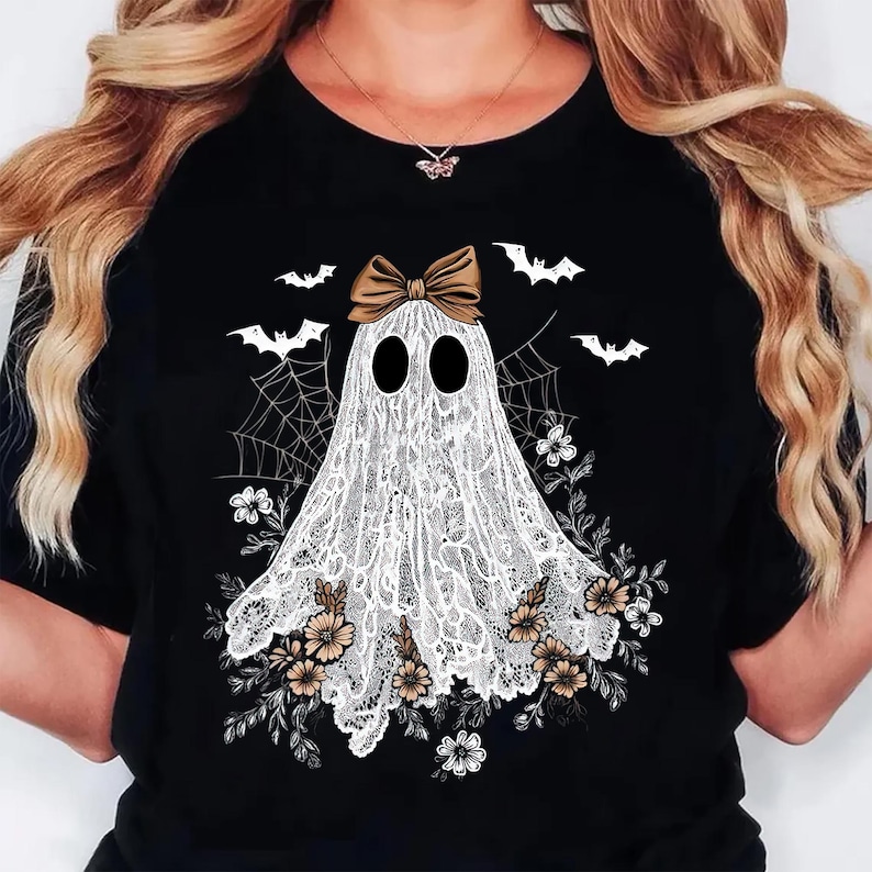 May include: Black t-shirt featuring a white ghost design with a brown bow, surrounded by bats, flowers, and a spiderweb. The ghost is made of lace. A Halloween-themed graphic tee.