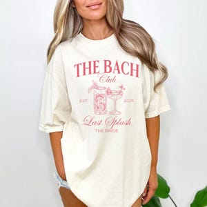 May include: A cream-coloured t-shirt featuring a pink graphic. The graphic includes the text "THE BACH Club", a cocktail illustration, and "Last Splash". The shirt also has "EST. 2025" and "THE BRIDE" printed on it.