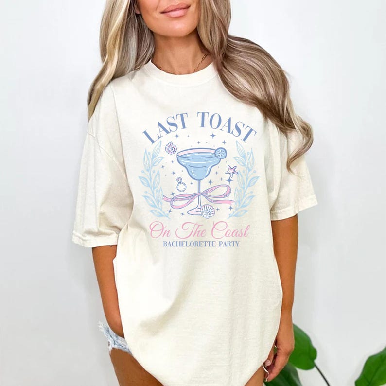 May include: Cream-colored t-shirt with a light blue and pink graphic. The graphic includes a cocktail, ring, and seashell, with the text "LAST TOAST" and "On The Coast Bachelorette Party".