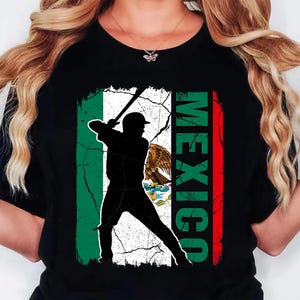 May include: Black t-shirt with a baseball player silhouette holding a bat, set against a distressed Mexican flag design. The word "MEXICO" is vertically printed in green and red. The shirt has a crew neck.