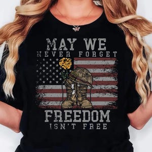 May include: A black t-shirt featuring a graphic of a military helmet resting on a pair of combat boots with a yellow flower. The design includes the American flag and the text MAY WE NEVER FORGET FREEDOM ISN'T FREE in a distressed grey font.