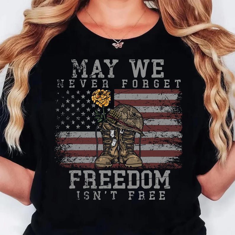 May include: A black t-shirt featuring a graphic of a military helmet resting on a pair of combat boots with a yellow flower. The design includes the American flag and the text MAY WE NEVER FORGET FREEDOM ISN'T FREE in a distressed grey font.
