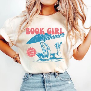 Peut inclure: T-shirt cr&egrave;me avec l'inscription "BOOK GIRL Summer" en rose et bleu. Le graphisme repr&eacute;sente une femme lisant sous un parasol de plage. Un petit badge rose indique "DIVE INTO A GOOD BOOK!"