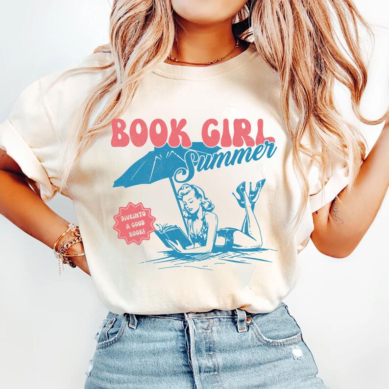 Peut inclure: T-shirt cr&egrave;me avec l'inscription "BOOK GIRL Summer" en rose et bleu. Le graphisme repr&eacute;sente une femme lisant sous un parasol de plage. Un petit badge rose indique "DIVE INTO A GOOD BOOK!"