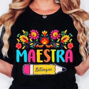 May include: Black t-shirt with a colorful floral design and the word "MAESTRA" in large, multi-colored letters. Below, a yellow pencil with the word "Bilingue" is featured. The design is vibrant and eye-catching.