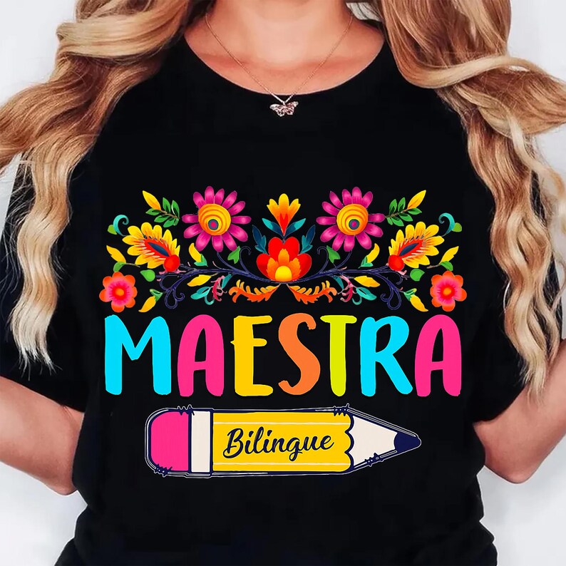 May include: Black t-shirt with a colorful floral design and the word "MAESTRA" in large, multi-colored letters. Below, a yellow pencil with the word "Bilingue" is featured. The design is vibrant and eye-catching.