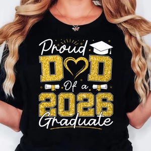 K&ouml;nnte beinhalten: Schwarzes T-Shirt mit goldglitzerndem Text "Proud Dad Of a 2026 Graduate". Das Design umfasst ein Herz, eine Abschlusskappe und Schriftrollen. Das Shirt ist eine Feier f&uuml;r einen Absolventen.