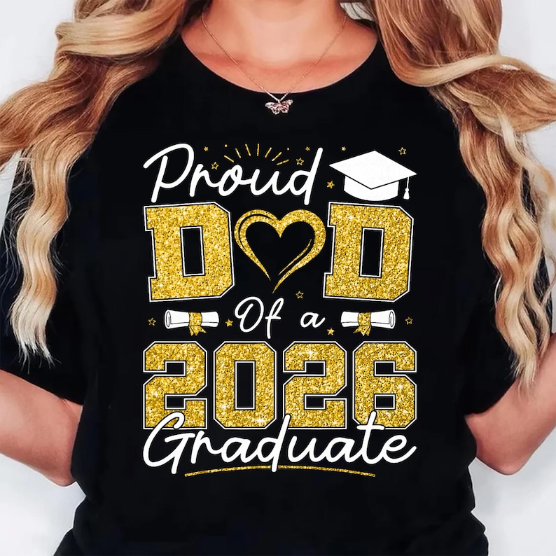 K&ouml;nnte beinhalten: Schwarzes T-Shirt mit goldglitzerndem Text "Proud Dad Of a 2026 Graduate". Das Design umfasst ein Herz, eine Abschlusskappe und Schriftrollen. Das Shirt ist eine Feier f&uuml;r einen Absolventen.