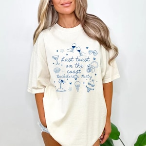 May include: Cream-colored t-shirt with a blue graphic design. The design includes the text "Last toast on the coast Bachelorette Party" along with illustrations of cocktails, sunglasses, and other beach-themed icons.