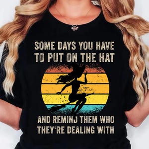 May include: Black t-shirt with the text "SOME DAYS YOU HAVE TO PUT ON THE HAT AND REMIND THEM WHO THEY'RE DEALING WITH." A silhouette of a witch on a broom is set against a yellow, orange, and blue sunset.