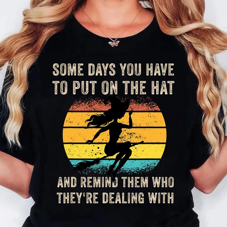 May include: Black t-shirt with the text "SOME DAYS YOU HAVE TO PUT ON THE HAT AND REMIND THEM WHO THEY'RE DEALING WITH." A silhouette of a witch on a broom is set against a yellow, orange, and blue sunset.