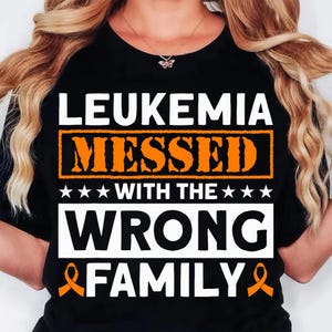 May include: Black t-shirt with the text "LEUKEMIA MESSED WITH THE WRONG FAMILY" in white and orange. The design includes orange awareness ribbons and stars. The shirt is a crew neck.
