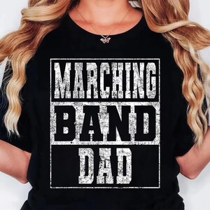 K&ouml;nnte beinhalten: Schwarzes T-Shirt mit dem Text "MARCHING BAND DAD" in verblichener wei&szlig;er Schrift. Der Text ist in drei horizontalen Zeilen innerhalb eines wei&szlig;en rechteckigen Rahmens angeordnet. Das T-Shirt hat einen Rundhalsausschnitt.