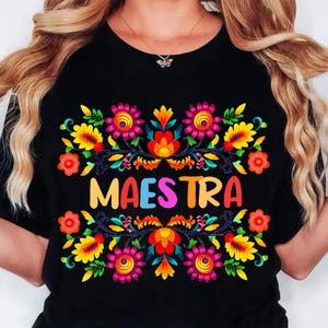 May include: Black t-shirt featuring the word "MAESTRA" in colorful letters, surrounded by a vibrant floral design. The design includes pink, yellow, orange, and blue flowers with green leaves, creating a festive look.