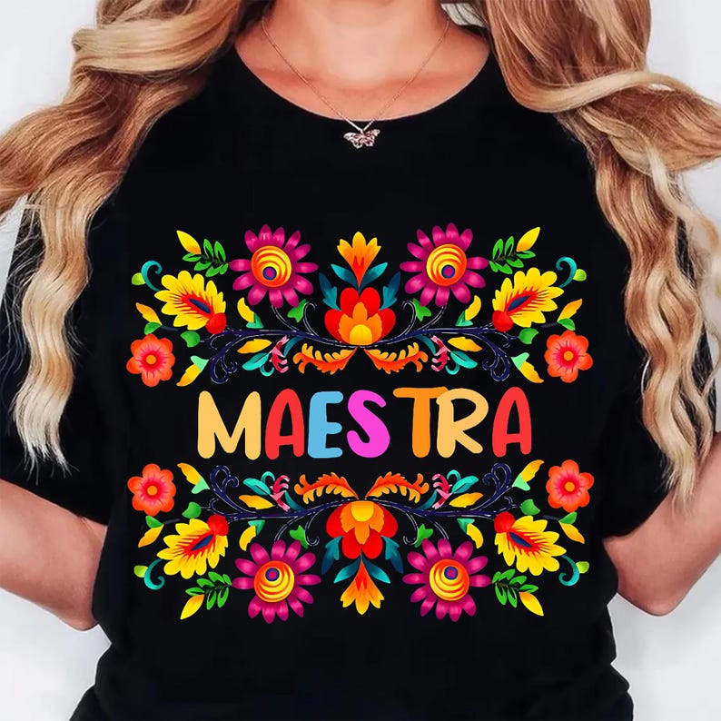 May include: Black t-shirt featuring the word "MAESTRA" in colorful letters, surrounded by a vibrant floral design. The design includes pink, yellow, orange, and blue flowers with green leaves, creating a festive look.