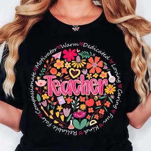 May include: Black t-shirt with a floral design surrounding the word "Teacher" in pink. The design includes flowers, hearts, and other elements, with words like "compassionate" and "loving" encircling the design.