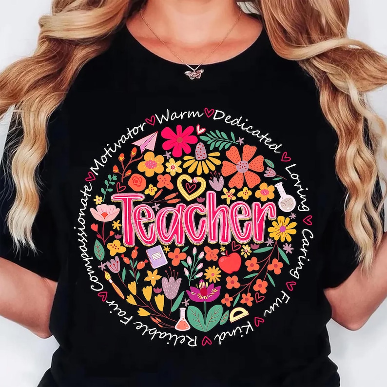 May include: Black t-shirt with a floral design surrounding the word "Teacher" in pink. The design includes flowers, hearts, and other elements, with words like "compassionate" and "loving" encircling the design.