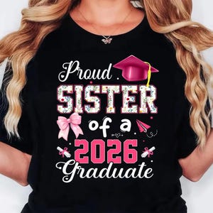 K&ouml;nnte beinhalten: Schwarzes T-Shirt mit dem Text "Proud SISTER of a 2026 Graduate". Das Wort "SISTER" ist mit einem floralen Muster gef&uuml;llt. Eine rosa Abschlusskappe und eine Schleife sind ebenfalls im Design enthalten.