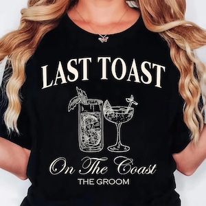 May include: Black t-shirt with the text "LAST TOAST" above a sketched illustration of two cocktails. Below the illustration, the text reads "On The Coast" and "THE GROOM". The design features a tall glass and a stemmed glass.
