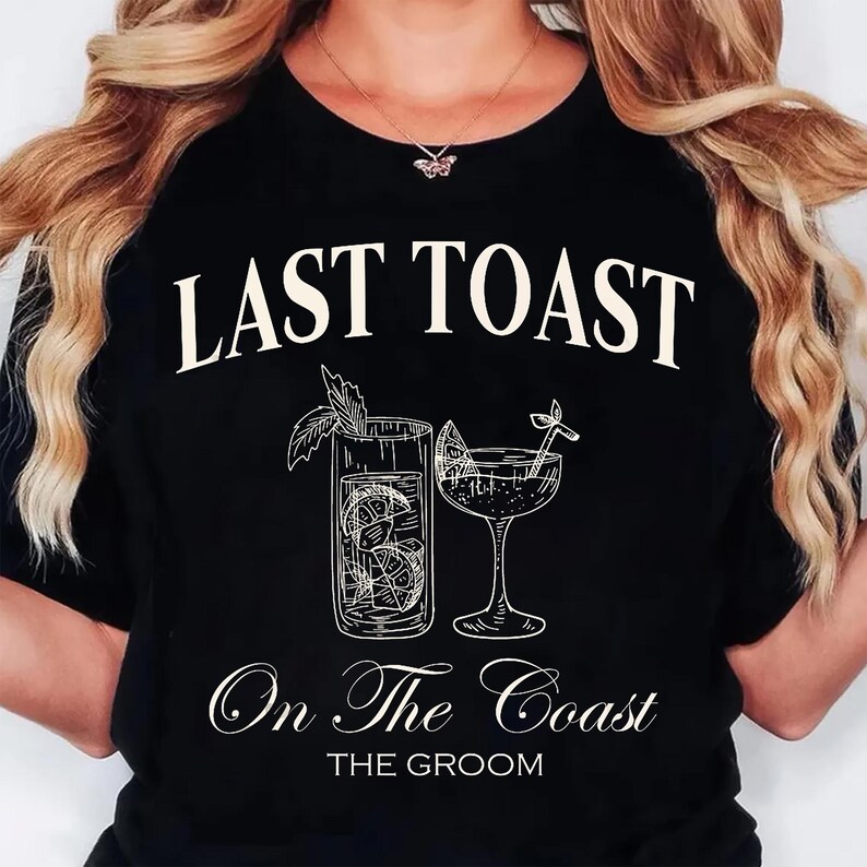 May include: Black t-shirt with the text "LAST TOAST" above a sketched illustration of two cocktails. Below the illustration, the text reads "On The Coast" and "THE GROOM". The design features a tall glass and a stemmed glass.