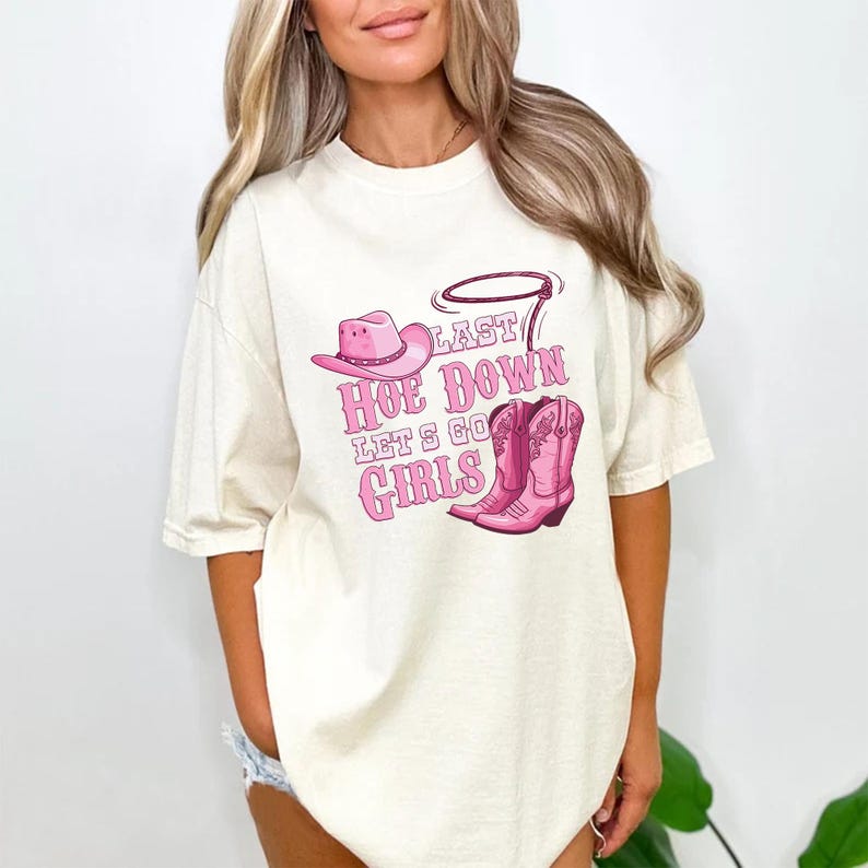 May include: Cream-colored t-shirt with a pink graphic. The graphic includes a cowboy hat, lasso, and cowboy boots, along with the text "Last Hoe Down Let's Go Girls."