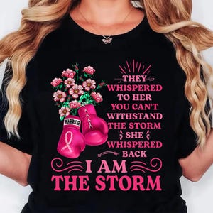 May include: Black t-shirt with a pink floral and boxing glove design. The design includes the text "THEY WHISPERED TO HER YOU CAN'T WITHSTAND THE STORM SHE WHISPERED BACK I AM THE STORM."