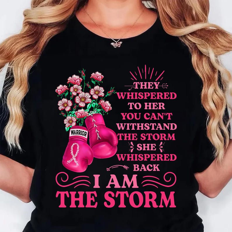 May include: Black t-shirt with a pink floral and boxing glove design. The design includes the text "THEY WHISPERED TO HER YOU CAN'T WITHSTAND THE STORM SHE WHISPERED BACK I AM THE STORM."