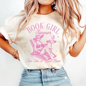 Peut inclure: T-shirt cr&egrave;me avec un motif rose. Le motif repr&eacute;sente une femme lisant dans une chaise longue, avec les mots "BOOK GIRL Summer" et "Dive Into A Good Book". Le design comprend un cocktail et un chapeau de soleil.