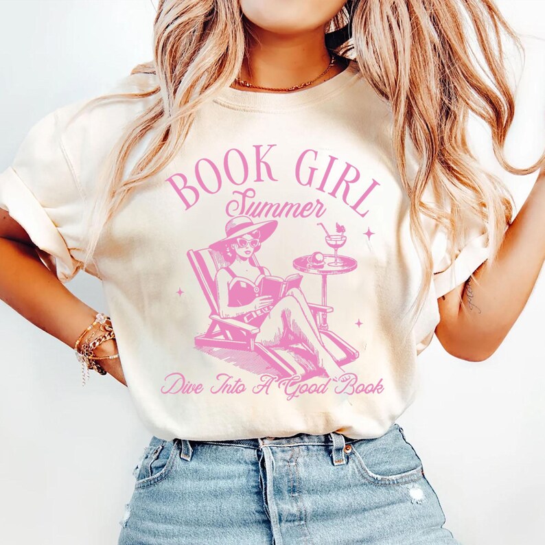 Peut inclure: T-shirt cr&egrave;me avec un motif rose. Le motif repr&eacute;sente une femme lisant dans une chaise longue, avec les mots "BOOK GIRL Summer" et "Dive Into A Good Book". Le design comprend un cocktail et un chapeau de soleil.