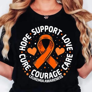 May include: Black t-shirt with an orange awareness ribbon, two orange hearts, and the words "HOPE, SUPPORT, LOVE, CARE, COURAGE, LEUKEMIA AWARENESS" in white. The design is circular and features white dots.